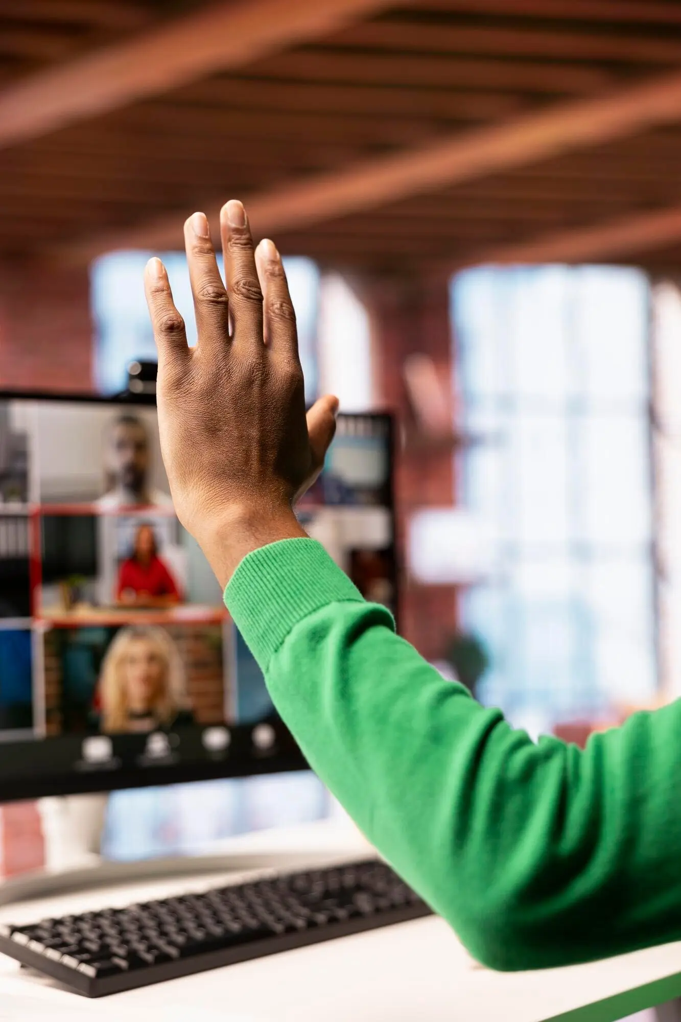 Remote worker at a home office desk raising a hand to speak during an online video call.